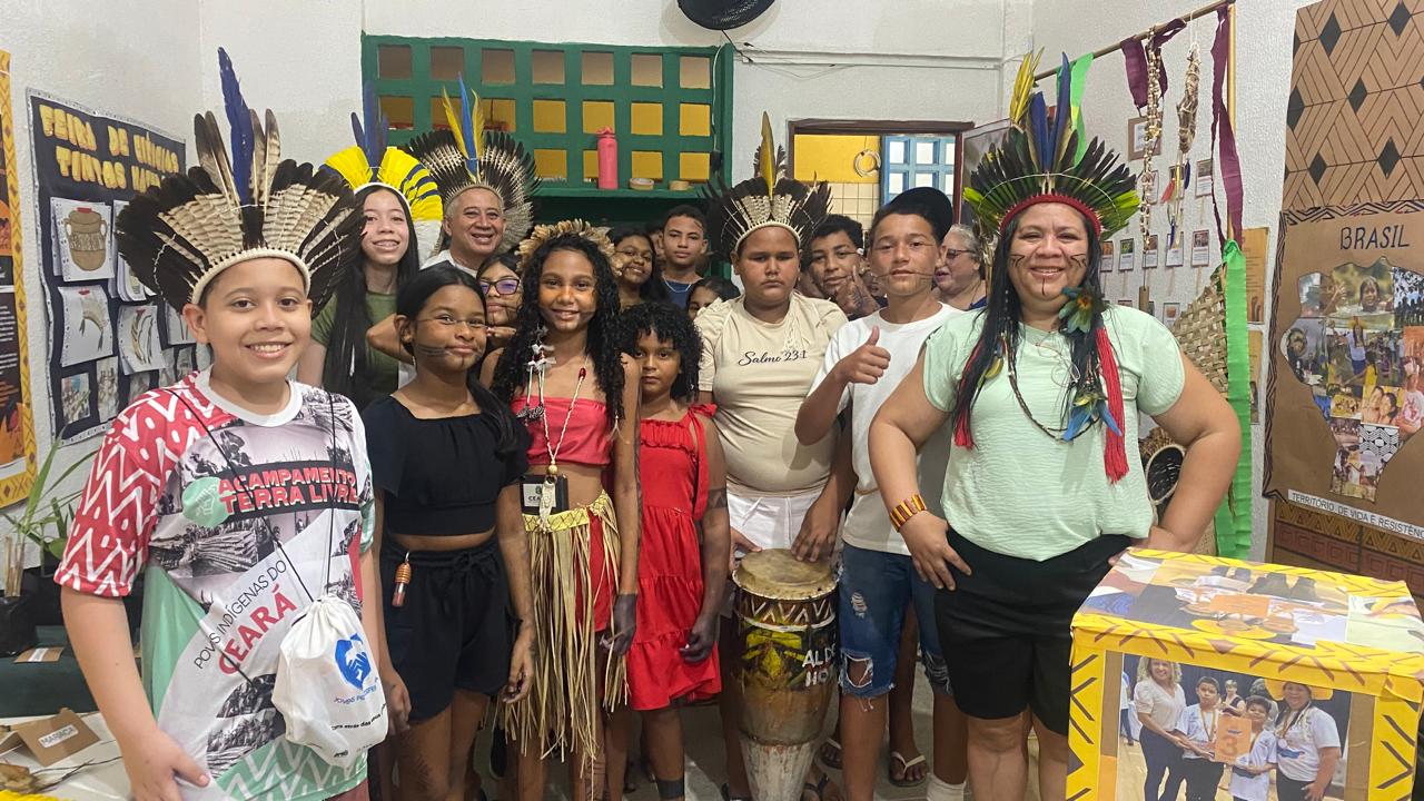 A fotografia mostra a professora e os estudantes em uma sala de aula usando adereços indígenas